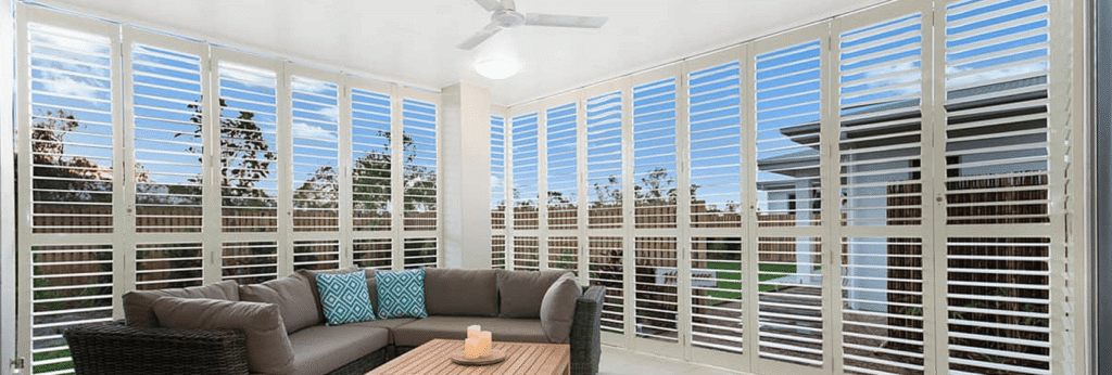 A modern sunroom with white plantation shutters, a sectional sofa with blue and patterned cushions, a wooden coffee table, and a ceiling fan.