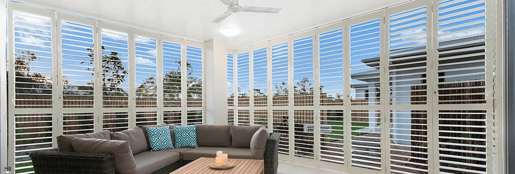 A modern sunroom with white plantation shutters, a sectional sofa with blue and patterned cushions, a wooden coffee table, and a ceiling fan.