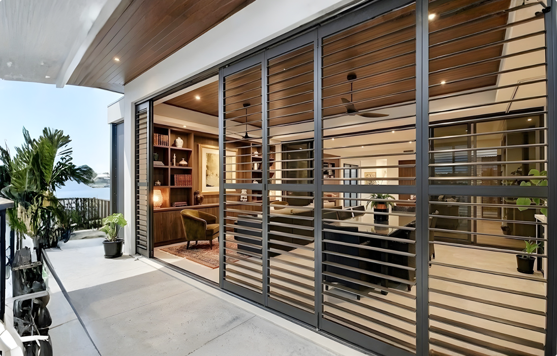 A modern living space with large glass sliding doors and horizontal metal slats, showing indoor plants, bookshelves, and seating, with a view of water and greenery outside.