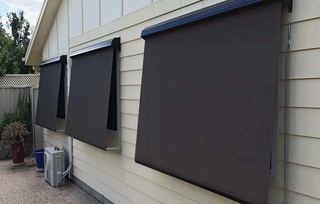 Three outdoor roller blinds are mounted on the exterior windows of a light-colored house, partially extended to provide shade.