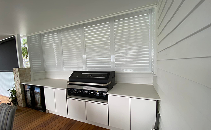 Outdoor kitchen with a built-in stainless steel barbecue grill, white cabinets, and white shutters behind on a covered patio with wooden flooring.