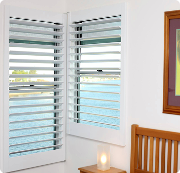 White plantation shutters cover two corner windows in a room with a water view. A wooden headboard, candle, and a framed picture are also visible.