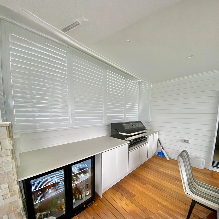Outdoor kitchen area with wooden flooring, a barbecue grill, white cabinets, beverage fridge, and white shuttered windows.