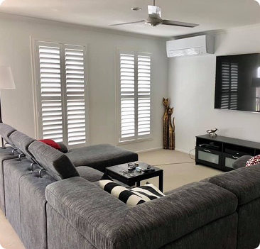 Modern living room with a gray sectional sofa, black TV stand, wall-mounted TV, decorative statues, and white shuttered windows.