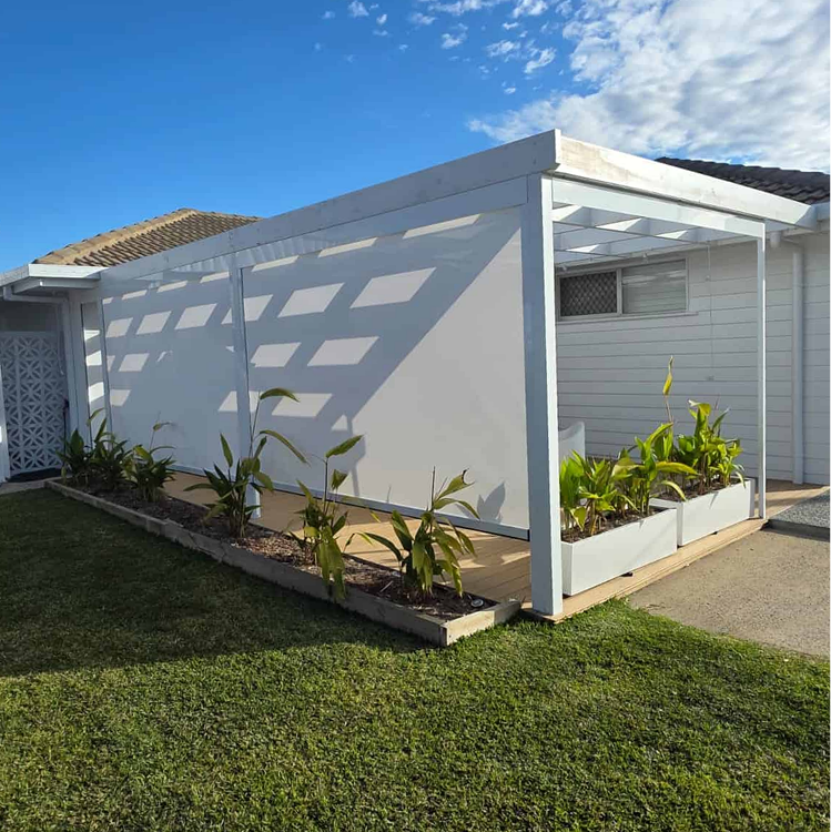 White pergola with partial roof, side privacy screen, and planter boxes with young tropical plants beside a house on a lawn.