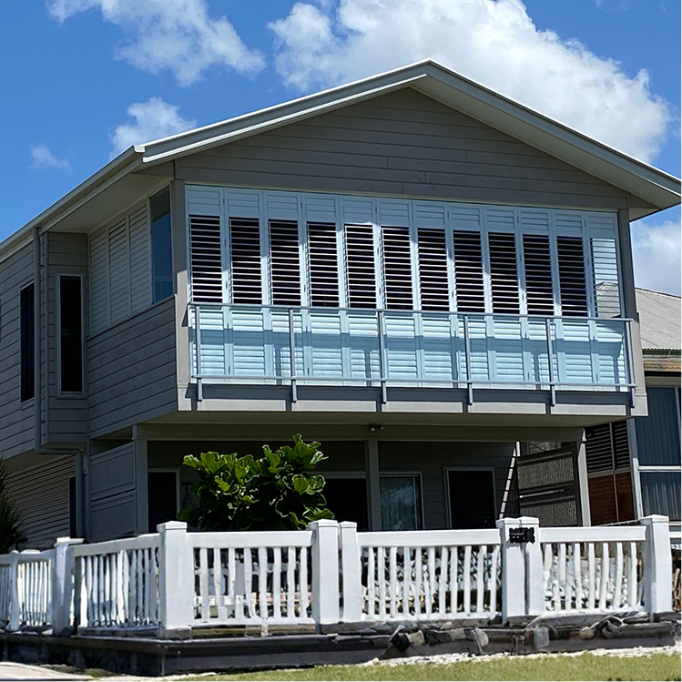 Two-story house with gray siding, large horizontal windows with shutters, a white balcony railing, and a small fenced yard. Blue sky with scattered clouds in the background.