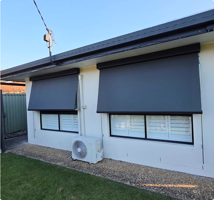 Two large, dark outdoor roller blinds are partially lowered over two windows on the exterior of a single-story house with a small air conditioning unit visible below.