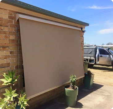 A beige outdoor roller blind is extended over a window on a brick wall, with potted plants placed nearby and a parked vehicle in the background.