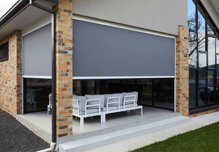 Outdoor patio with brick columns, glass walls, and a grey retractable screen. White outdoor furniture is arranged on the concrete patio floor.