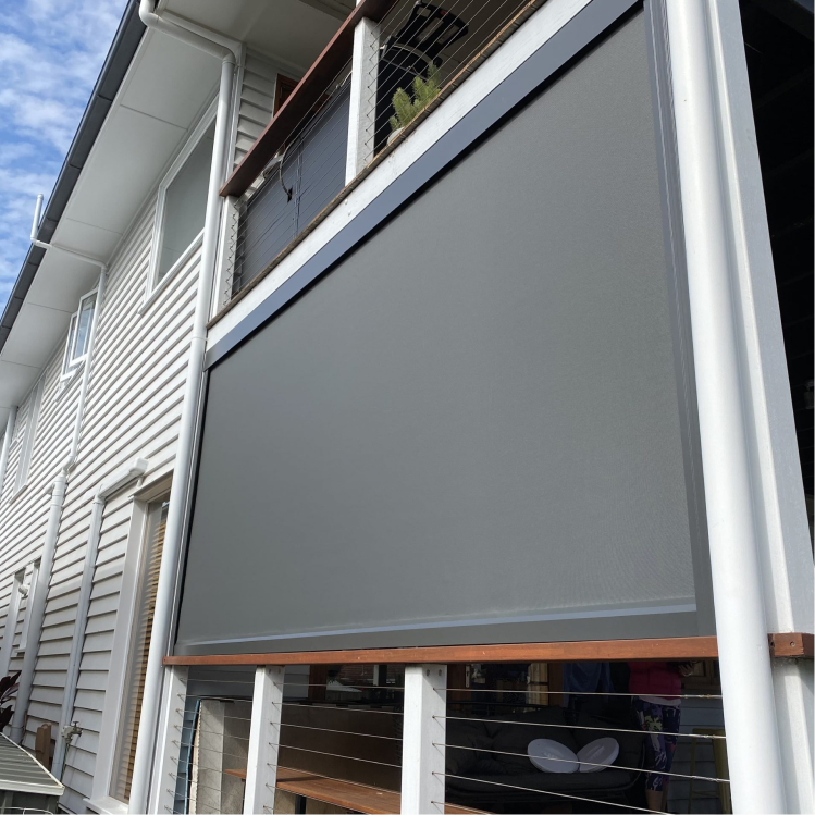 A gray outdoor roller blind covers a balcony area on a two-story white house with horizontal siding.