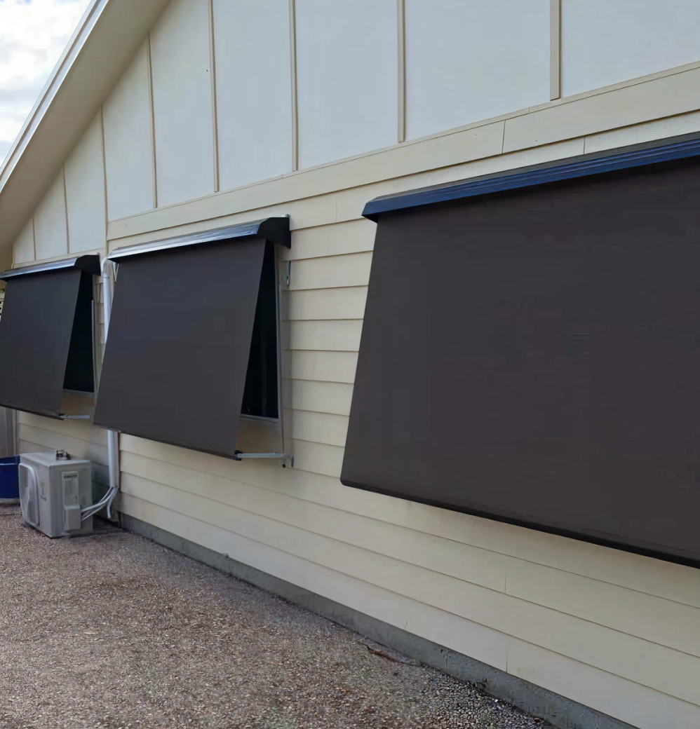 awnings Three large, dark-colored awnings are extended over windows on the side of a light-colored building with horizontal siding and a gravel ground.