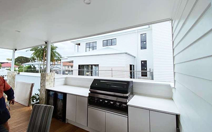 Outdoor kitchen area with a built-in gas grill, white countertops, and cabinets, overlooking a neighboring white house; part of a deck and chairs are visible.