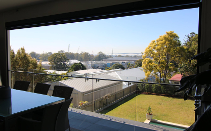 View from a covered balcony showing a backyard, patio, glass railing, and a neighborhood with trees and buildings under a clear sky.