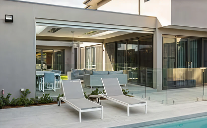 Two white lounge chairs beside a pool in front of a modern house with large glass windows and a visible indoor seating area.