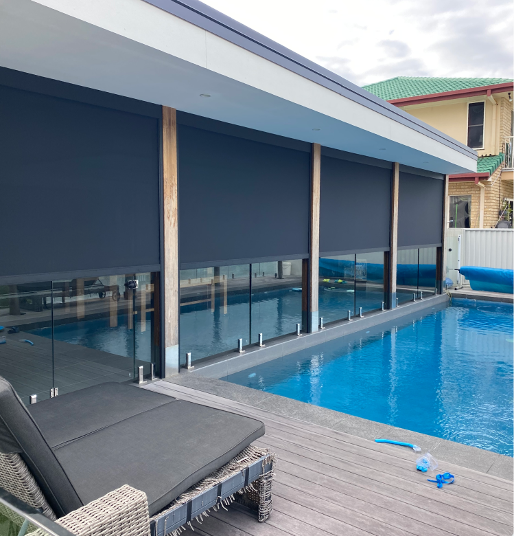 heavy A modern outdoor pool next to a building with large, dark roller shades covering the windows. A lounge chair sits on the wooden deck in the foreground.