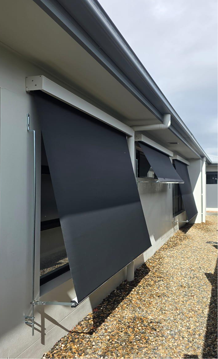 Exterior view of a modern building with two black retractable awnings partially extended over windows, above a gravel pathway.