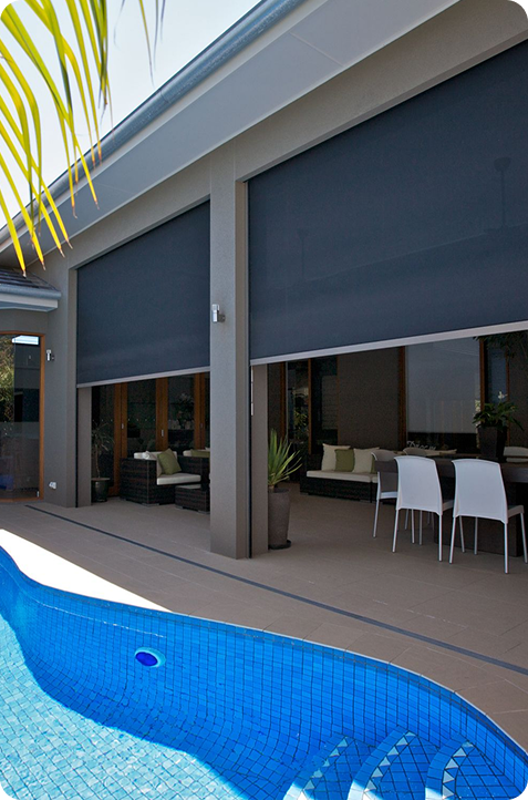 A modern patio with black roller blinds partially down, outdoor seating area, dining table, and a blue-tiled swimming pool in the foreground.