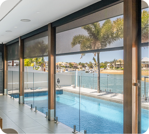 Indoor pool area with glass barriers, shaded by large retractable blinds, overlooking a waterfront with boats and palm trees outside.