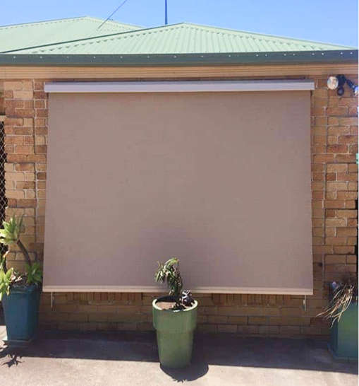 A beige outdoor roller blind covers a window on a brick house. Two green potted plants are positioned in front of the blind on a concrete surface.