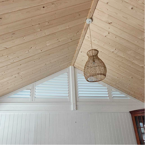 A slanted wooden ceiling with a wicker pendant light hanging and white horizontal blinds covering triangular windows.