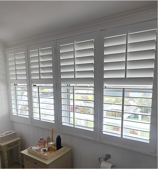 White plantation shutters cover a set of four windows in a bright room with toiletries and a toilet paper holder visible below the windowsill.
