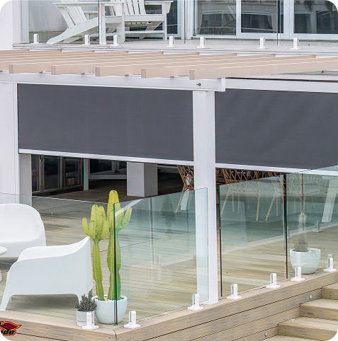 Outdoor pergola with a retractable gray shade, surrounded by glass panels, white chairs, and a potted cactus on a wooden deck.