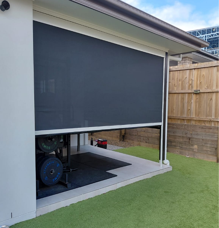 motirised Outdoor home gym area partially enclosed with a grey roller shade, visible gym weights, artificial grass, and a wooden fence in the background.