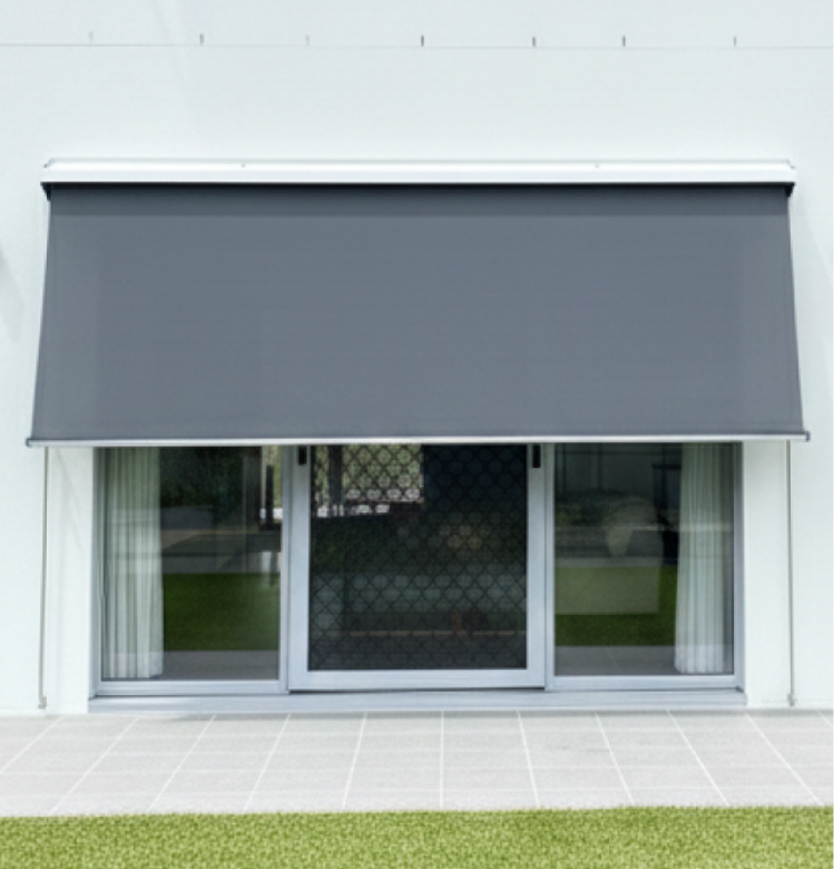 roller A dark grey outdoor awning covers a large window with white curtains, above a tiled patio and a strip of green grass.