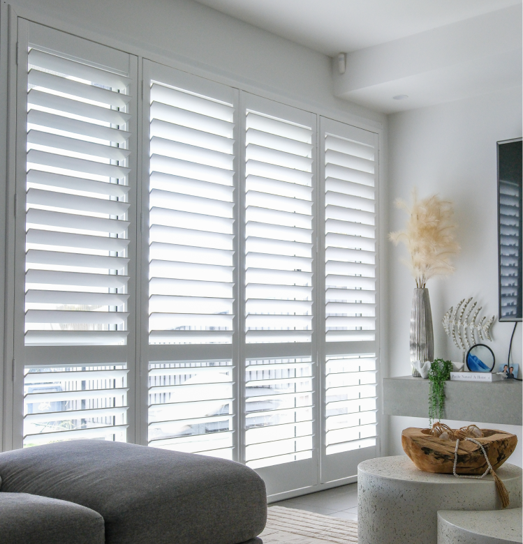 shuter Bright living room with large white plantation shutters, a grey sofa, round tables, a wooden bowl, and decorative items including a vase with pampas grass and a shell-shaped ornament.