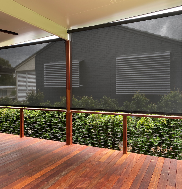 straght drop A covered wooden deck with metal railing, mesh roller blinds partially down, and view of neighboring building with louvered windows and green bushes.