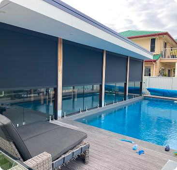 A modern backyard pool with glass fencing, covered patio with dark roller blinds, and poolside lounge chairs.