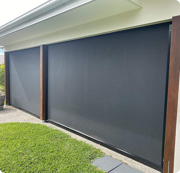 Two large black outdoor roller blinds cover a patio area, framed by wooden posts, with grass and paving stones in the foreground.