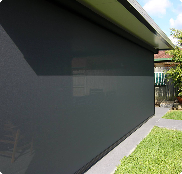 A large, dark outdoor roller blind fully extended on a patio, partially shading the area while grass and a house are visible nearby.