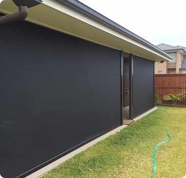 A backyard patio with black outdoor blinds fully lowered, covering the open sides of the patio area. A green garden hose lies on the grass nearby.