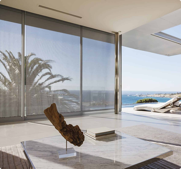 Modern living room with a marble coffee table, sculpture, and books, featuring floor-to-ceiling windows overlooking the ocean and palm trees.