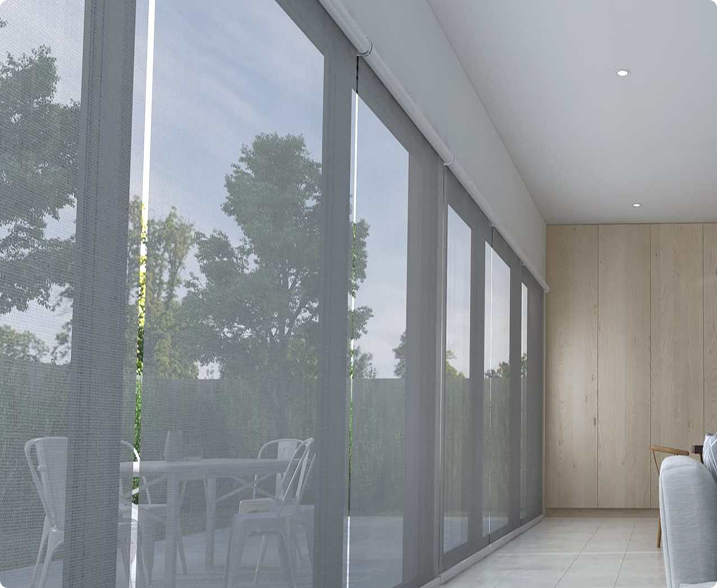 Large glass sliding doors with gray mesh blinds partially drawn, overlooking a patio with metal chairs and table, and trees visible outside. Indoor space has light-colored tiled floor and wooden wall.