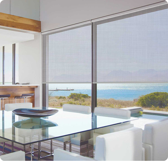 Modern dining room with a glass table and white chairs, large windows with shades, and a view of water and greenery outside.