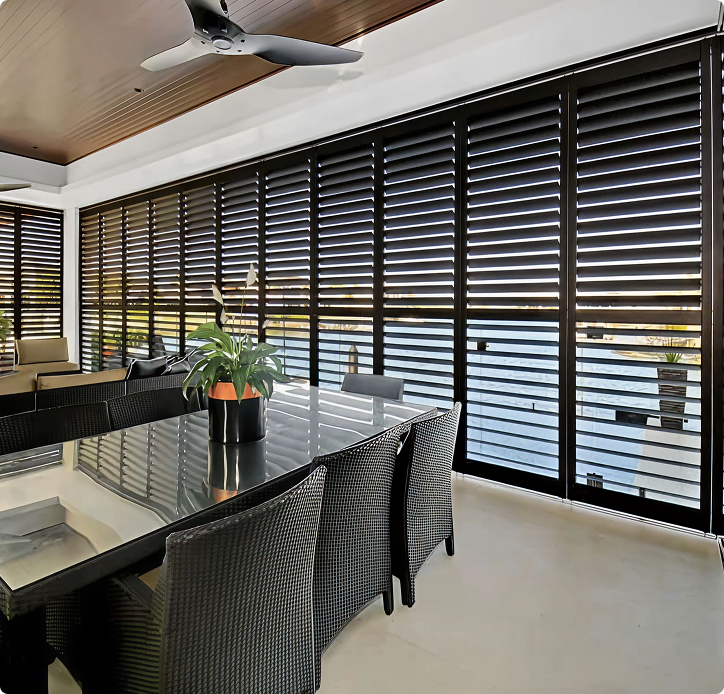 Modern dining area with a glass table, six wicker chairs, and a potted plant, next to large windows covered by horizontal black blinds.