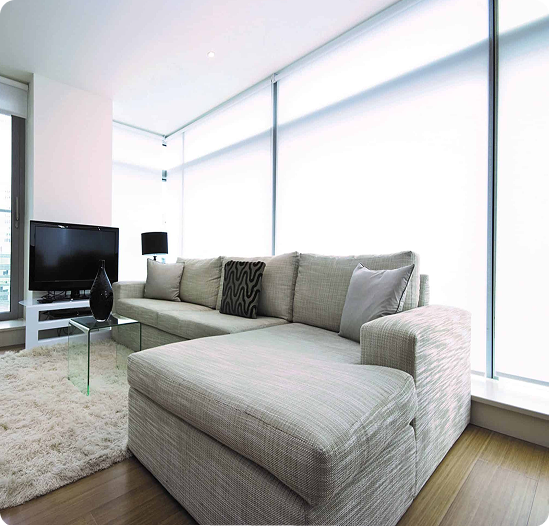 Modern living room with a light gray sectional sofa, glass coffee table, TV on a stand, and large windows with white blinds.