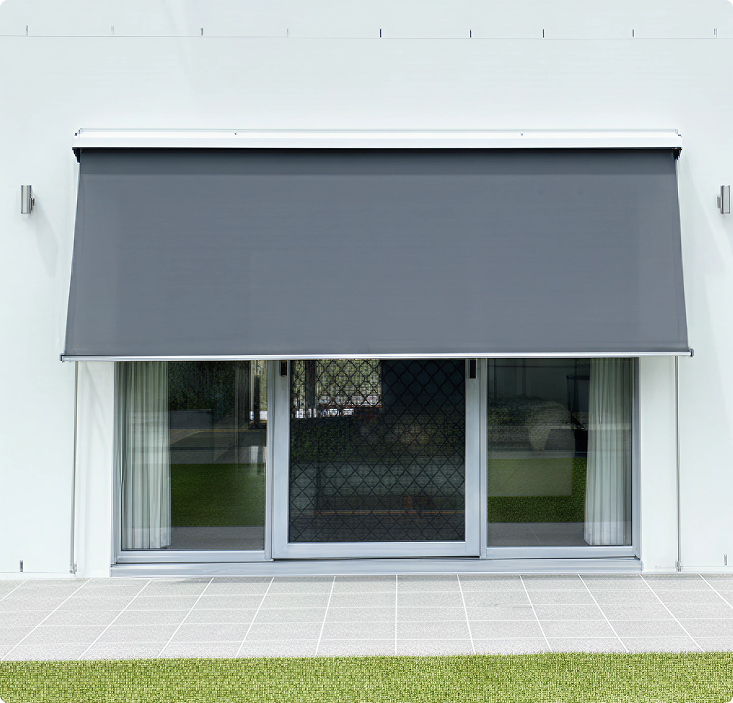 A gray outdoor roller blind is partially lowered over a sliding glass door with white curtains, opening onto a tiled patio and lawn.