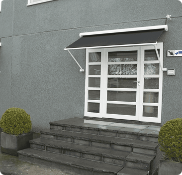 White glass double door with black awning above, set in a gray building. There are five stone steps and two round potted shrubs on either side of the entrance.
