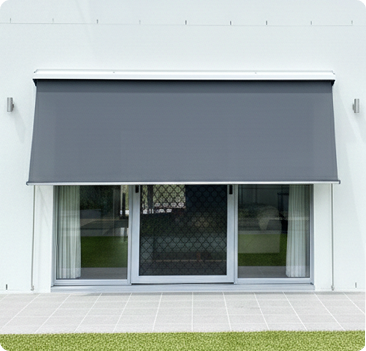 A gray outdoor window awning is extended over a large glass door with white curtains, set against a white wall with tiled flooring and grass in front.