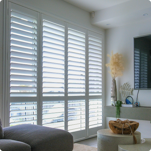 Large white plantation shutters cover floor-to-ceiling windows in a bright living room with modern decor, including a grey sofa and a TV mounted on the wall.