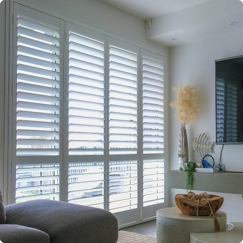 Modern living room with large white plantation shutters, a grey sofa, a TV on the wall, and decorative items on a corner console table.