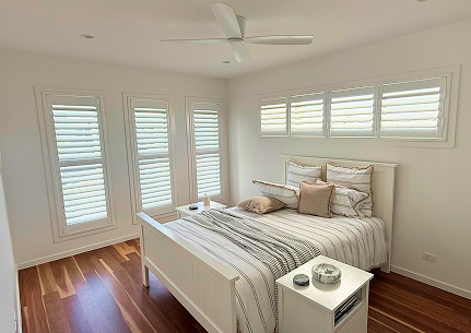 A bright bedroom with wooden floors, white walls, a ceiling fan, a white bed with striped bedding, and white shutters on the windows.