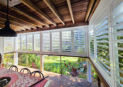 Covered outdoor patio with a wooden ceiling, white shutters, and a dining table with chairs, overlooking a green garden with plants and trees.