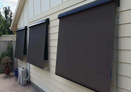 Three large, dark exterior roller blinds are installed on the windows of a light-colored house with a paved walkway and a garden area visible.
