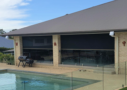 A covered patio with large black outdoor blinds, a barbecue grill, and a glass pool fence next to a swimming pool.