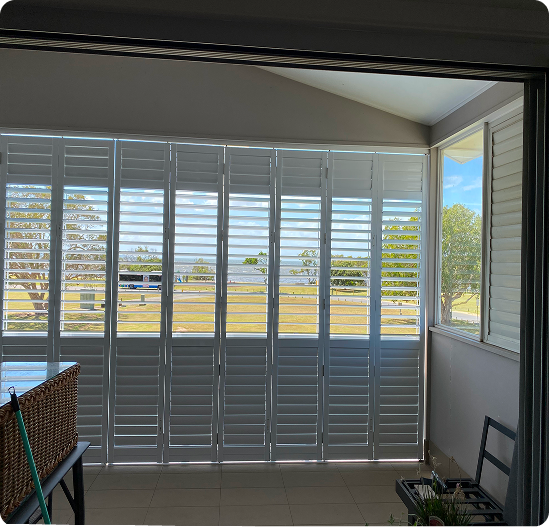 White plantation shutters cover floor-to-ceiling windows, partially open to show a grassy outdoor area with trees under a blue sky. A wicker basket and plant are in the room.