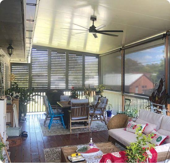 Covered patio with ceiling fan, table and chairs, potted plants, a sofa with cushions, and screened siding; natural light enters through slatted panels and mesh curtains.